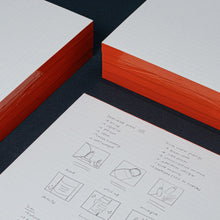 Load image into Gallery viewer, Two red-bound books on a black surface with a sheet of paper featuring illustrations and text.

